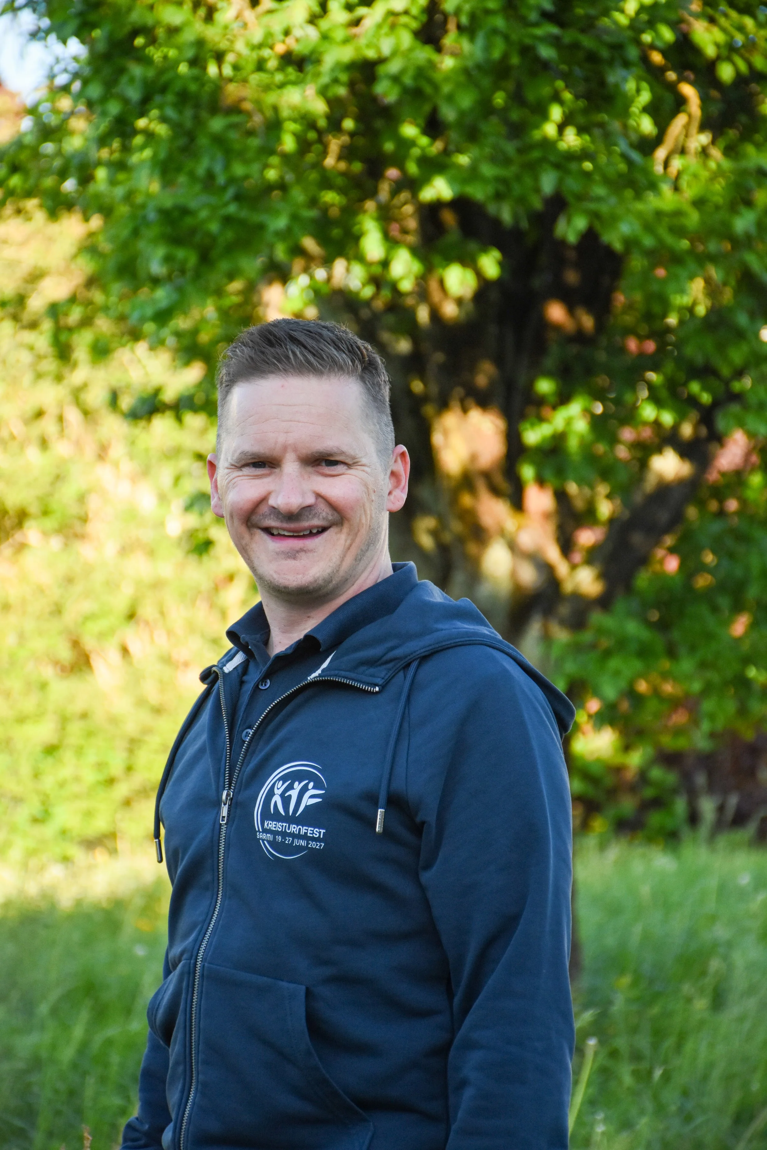 Ein lachender Mann trägt eine dunkelblaue Jacke mit einem Logo, im Hintergrund Bäume mit grünen Blättern, wahrscheinlich im Park, bei Tageslicht.