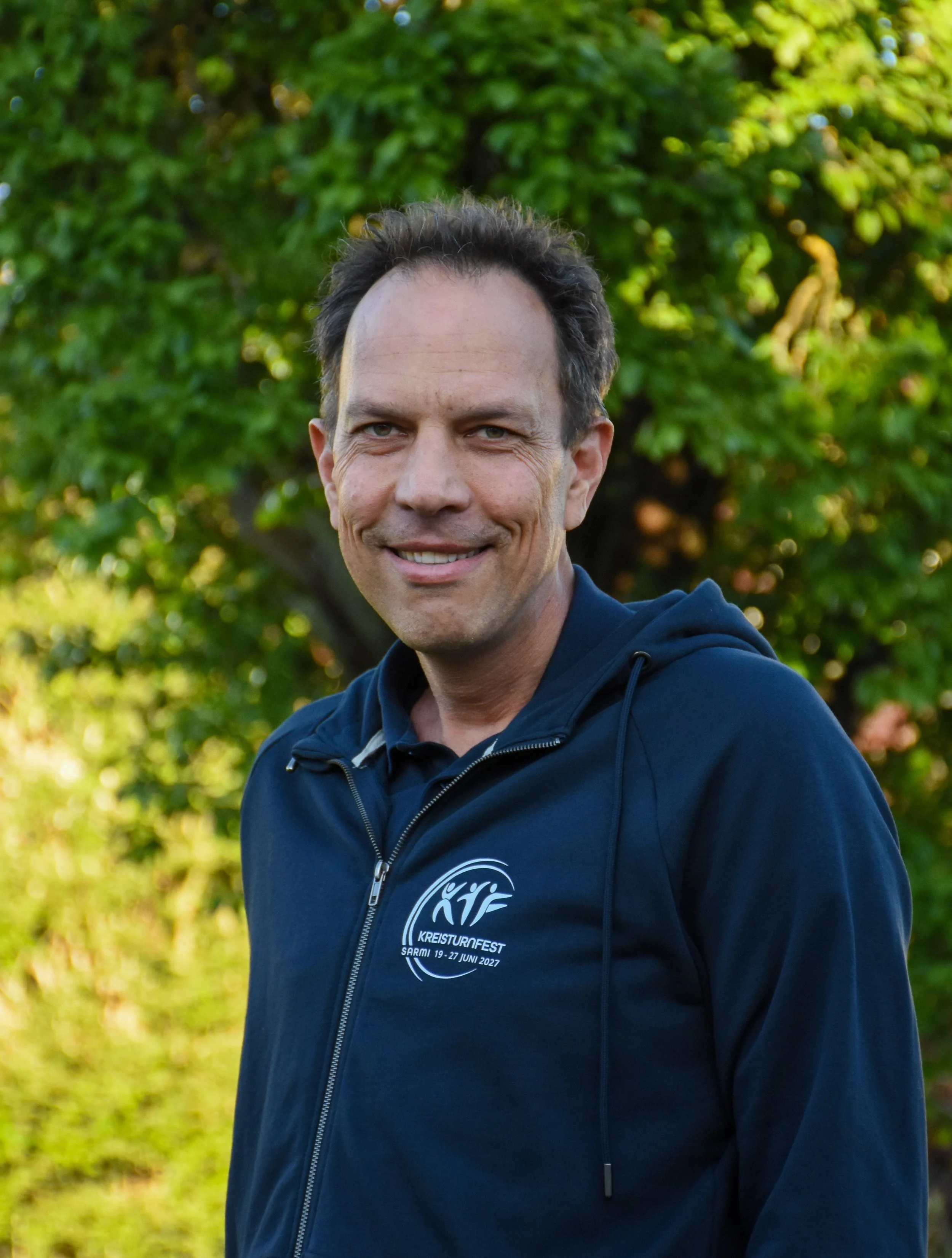 Ein lachender Mann trägt eine dunkelblaue Jacke mit einem Logo und Text, steht vor einem grünen Baum im Freien.