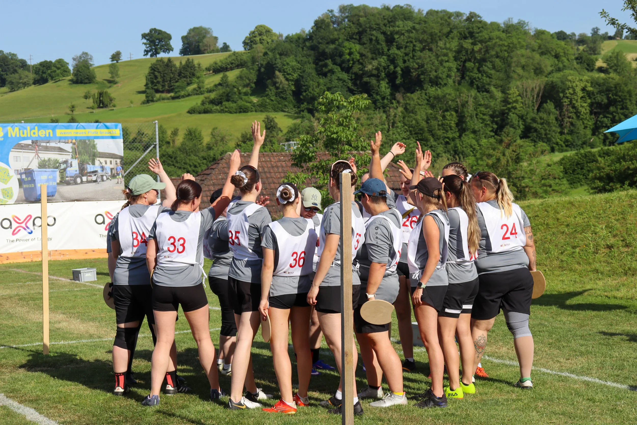 Gruppen von Frauen in Sportkleidung, die sich auf einem Rasenplatz für eine Sportveranstaltung versammeln, mit einer hügeligen Landschaft und Bäumen im Hintergrund.