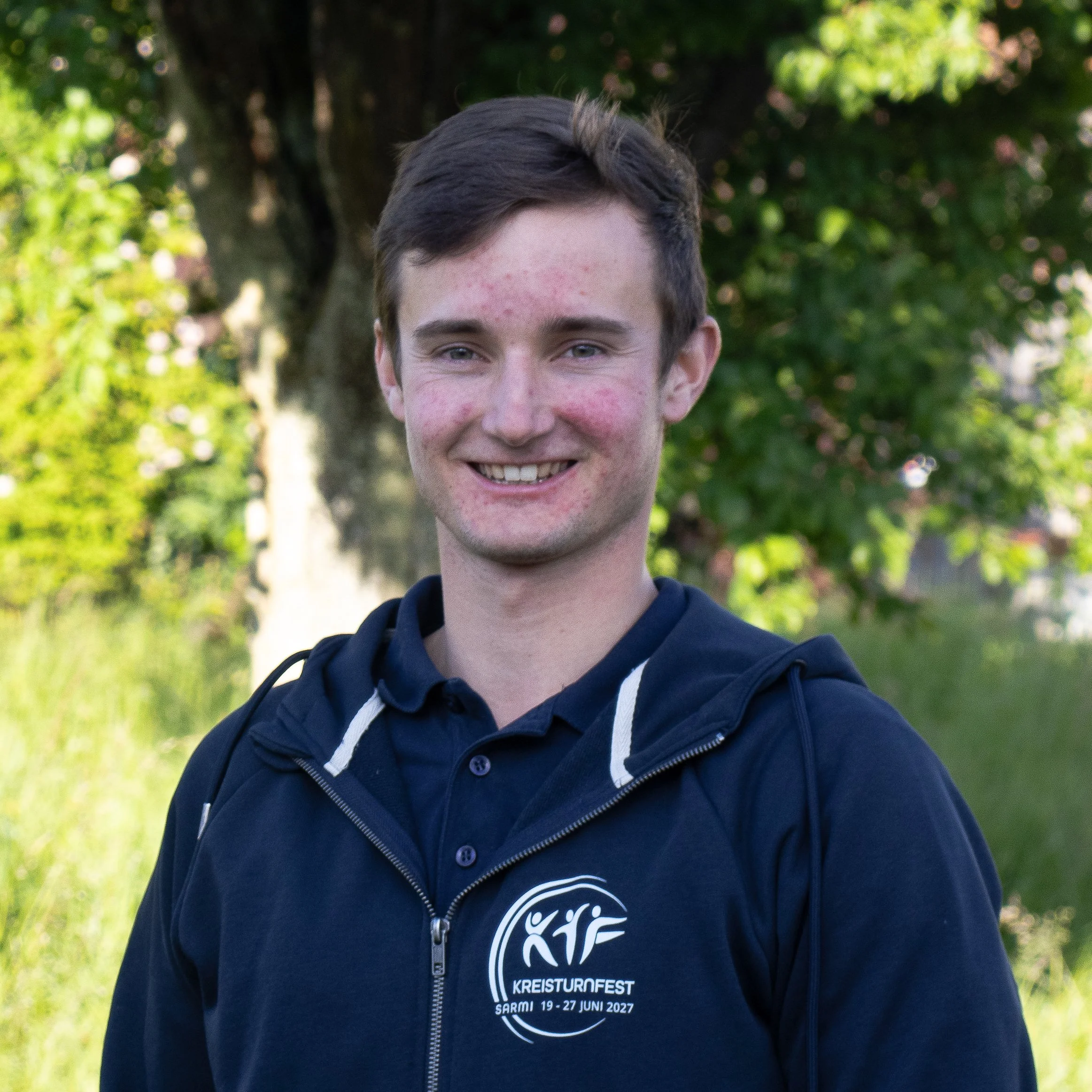 Junge Person trägt eine dunkelblaue Jacke und ein T-Shirt mit dem Text 'KREISTURNOFEST SARM1 19-27 Juni 2027' vor einem grünen Baum im Freien.