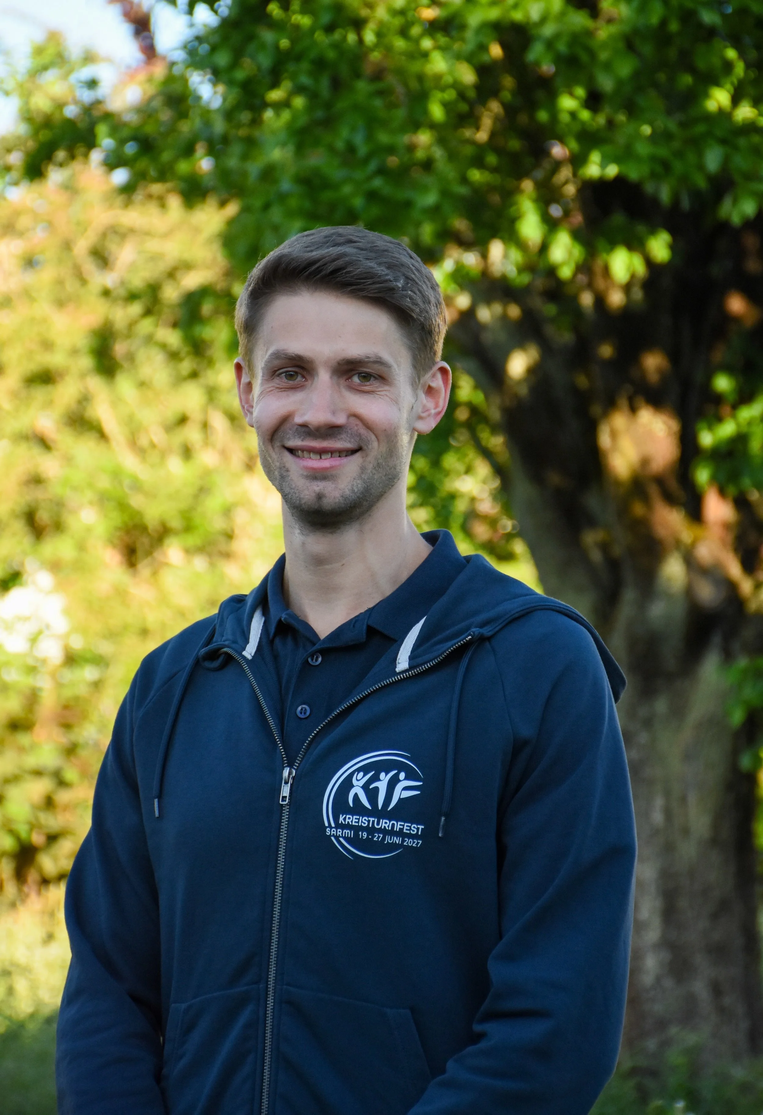 Ein junger Mann trägt eine dunkelblaue Jacke mit einem Logo und Text für den Kreis-Turnfest in einer grünen Parklandschaft bei sonnigem Wetter.