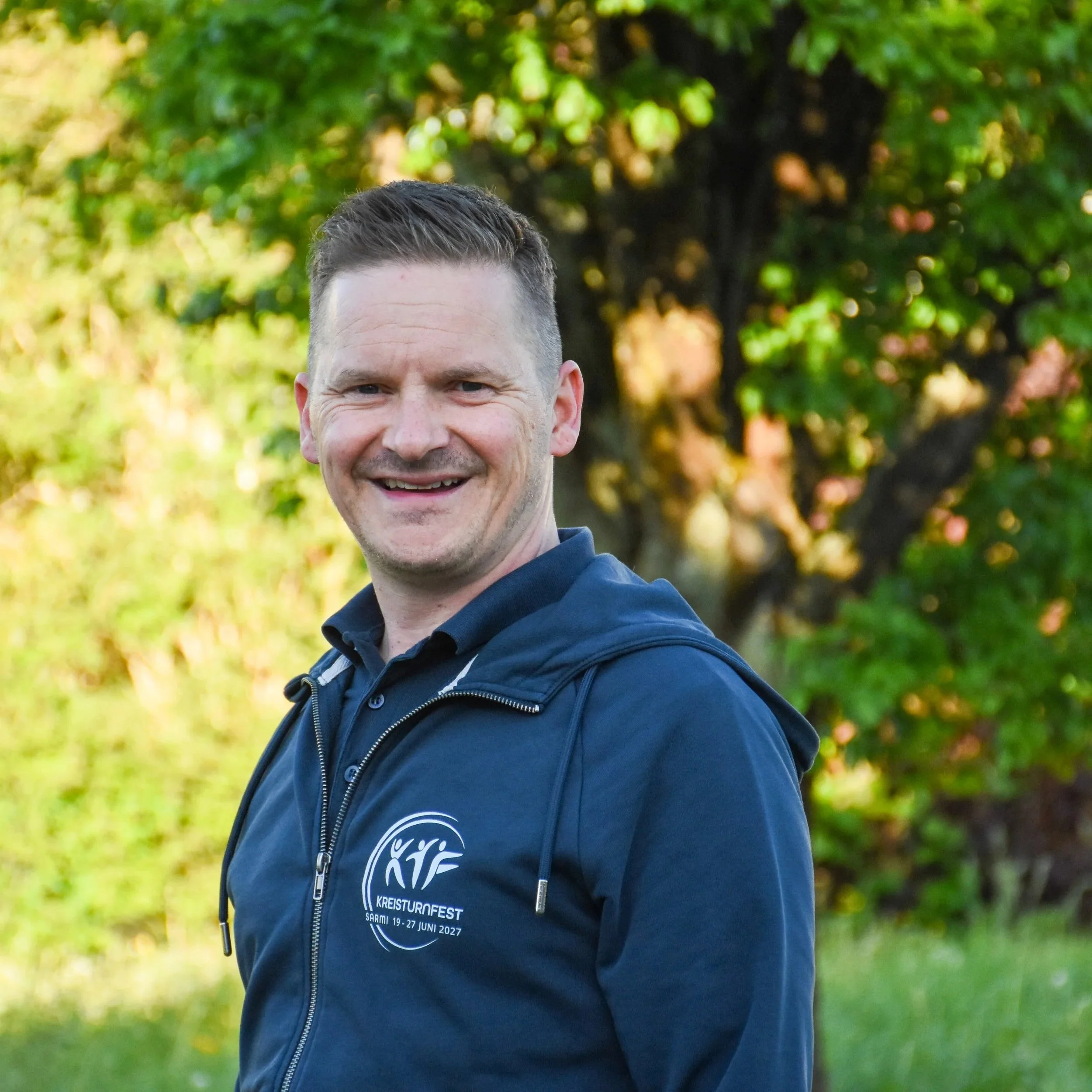 Lächelnder Mann trägt eine schwarze Jacke mit einem Kreislogo und steht vor einem Baum im Freien bei sonnigem Wetter.