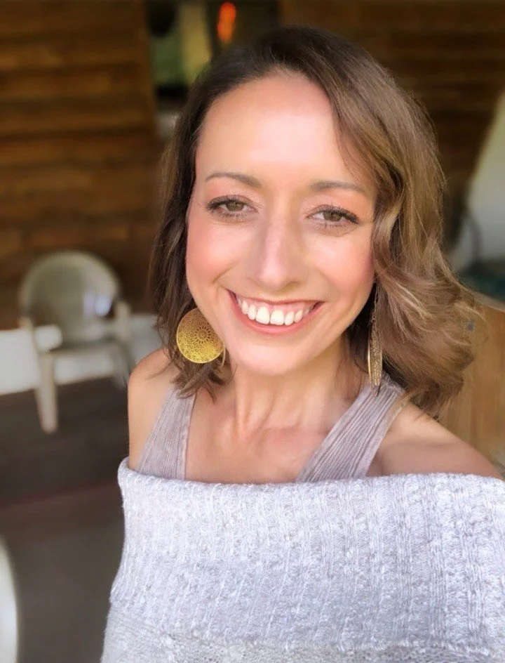 A smiling woman with shoulder-length wavy brown hair wearing yellow earrings and a light gray off-the-shoulder top, posing outdoors with a wooden background and blurred outdoor furniture.
