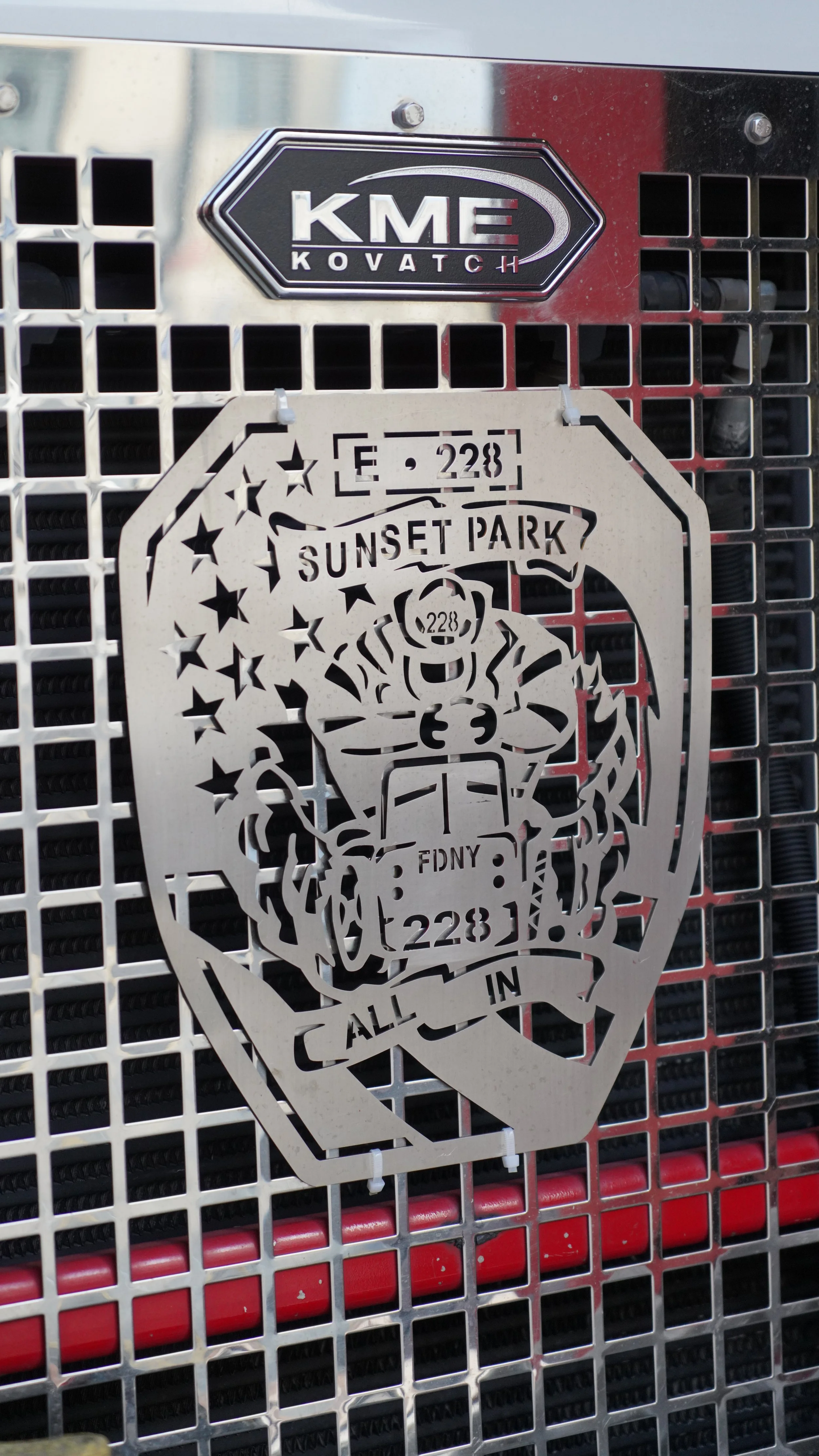 Close-up of a metal firefighter rescue tool with cut-out firefighter design, mounted on a metallic grid surface. There are also two badges or signage plaques above it.
