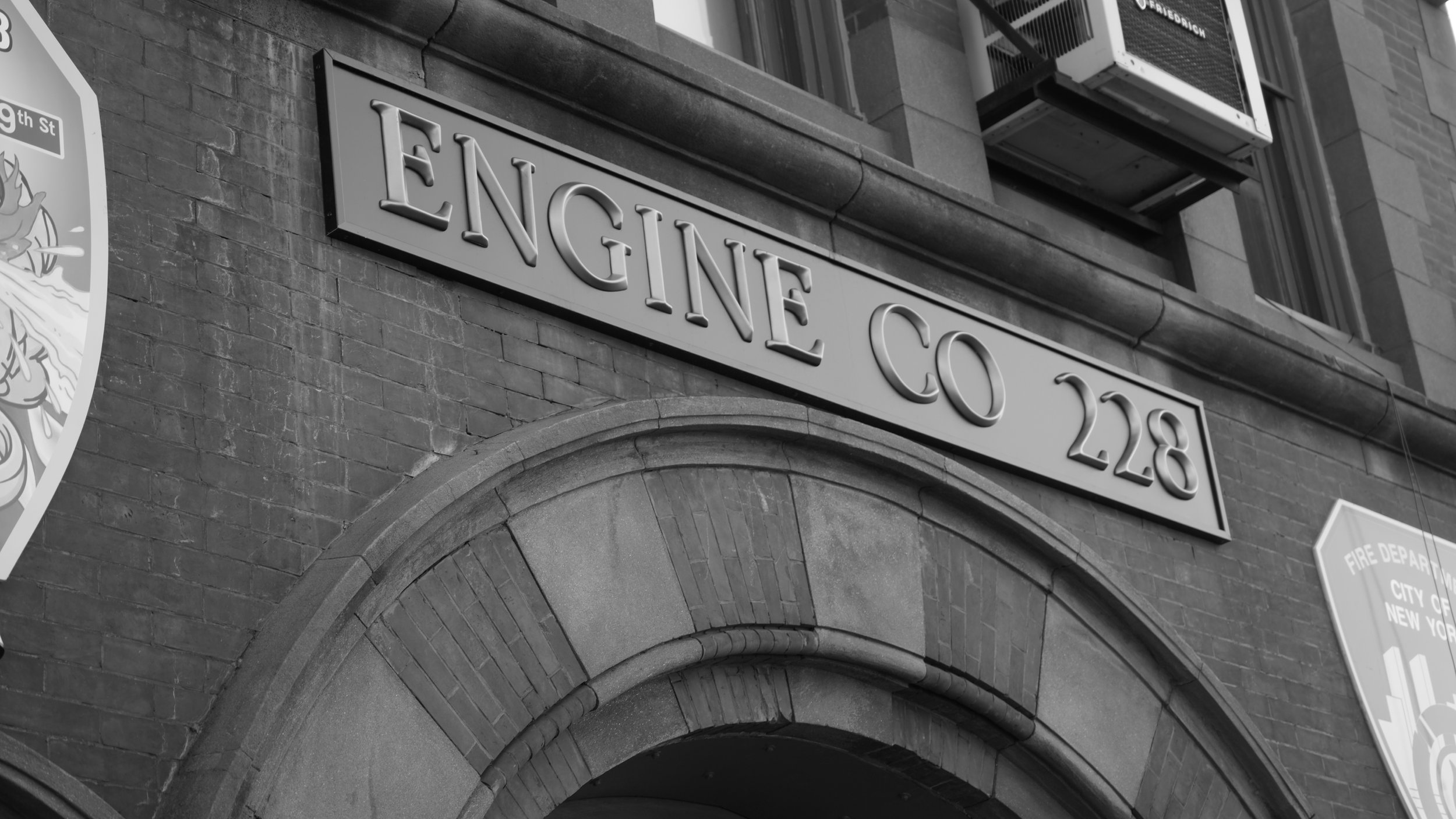 Black and white photo of a building with a sign reading "Engine Co 28" and an arched entrance made of brick and stone, with an air conditioning unit mounted above and a partial view of a shield-shaped emblem on the left.