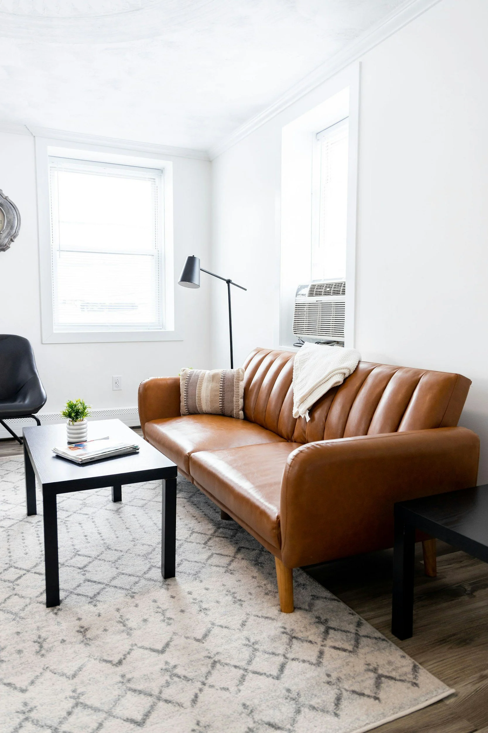 Living room with a tan leather sofa, black coffee table, black armchair, white walls, two large windows with blinds, a black floor lamp, a wall clock, and a patterned beige area rug.