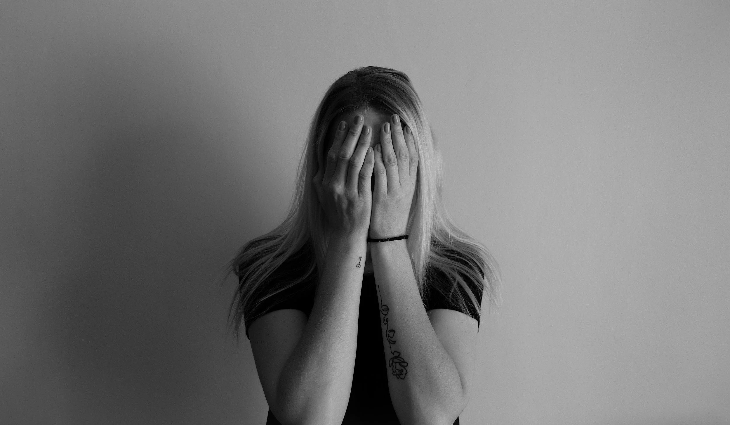 A woman with long hair covering her face with her hands, tattoos on her arms, and wearing a black shirt, standing against a plain wall in a black and white photograph.