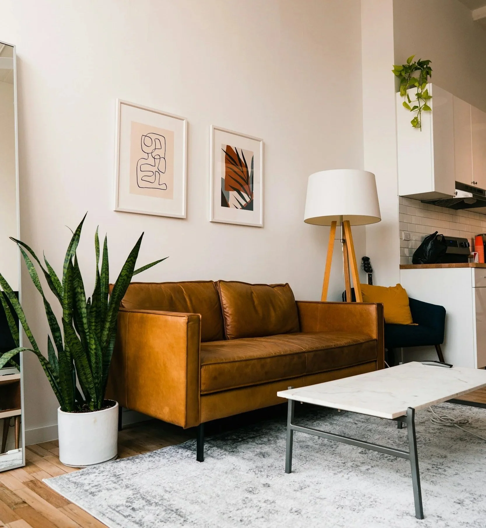Living room with a leather sofa, a large potted snake plant, two framed abstract art pieces on a white wall, a tripod floor lamp, a white marble coffee table, and a dining area in the background with white cabinets and a small countertop.