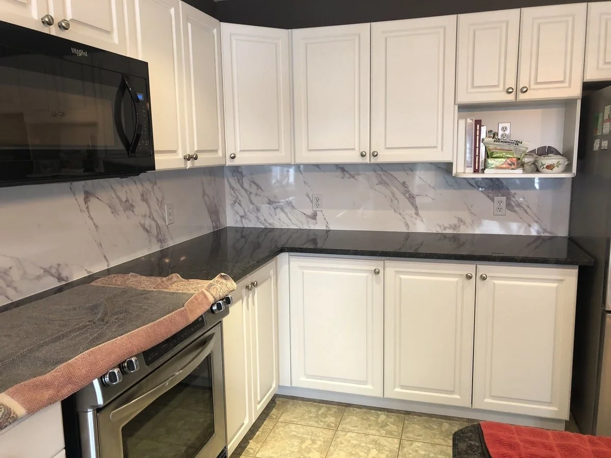 Kitchen with white cabinets, black marble countertops, marble backsplash, stainless steel microwave, oven, and refrigerator. Open shelf with books and kitchen items, tiled floor, and red towel on counter.