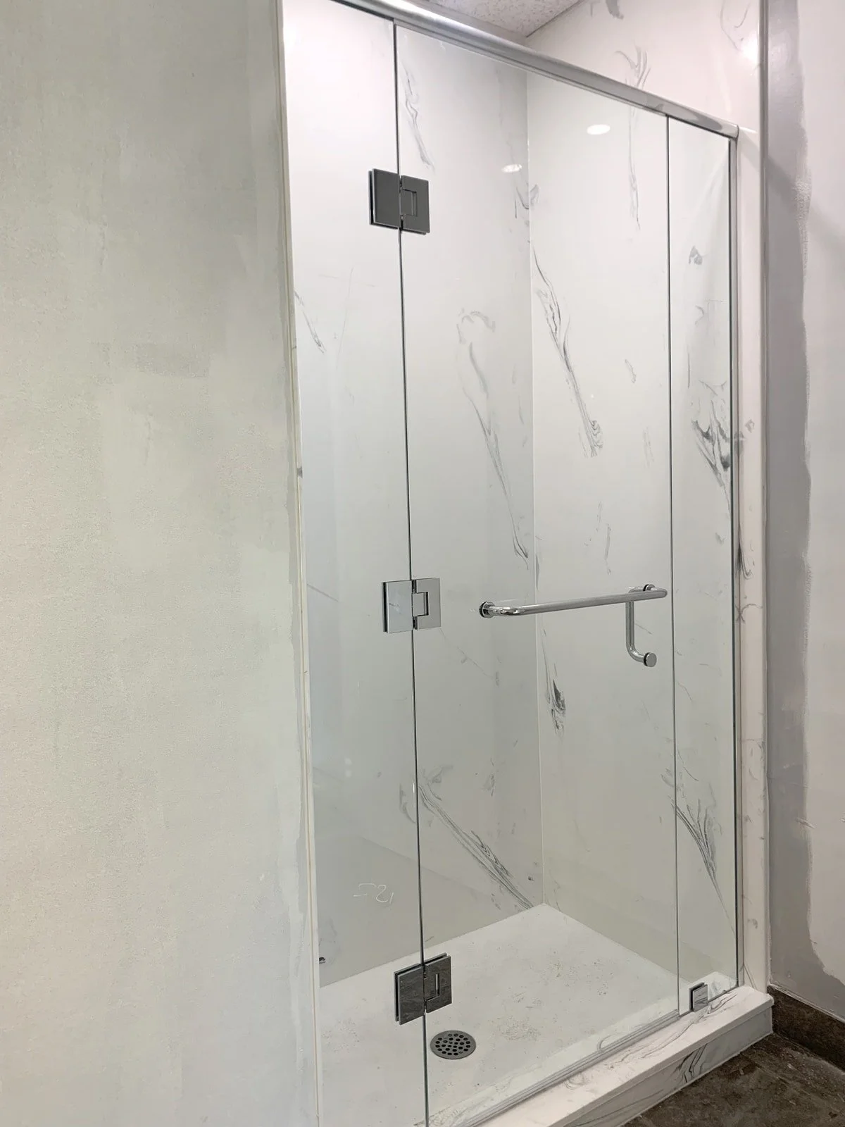 A modern glass shower enclosure with marble-patterned walls and a grab bar inside, with a drain on the floor.