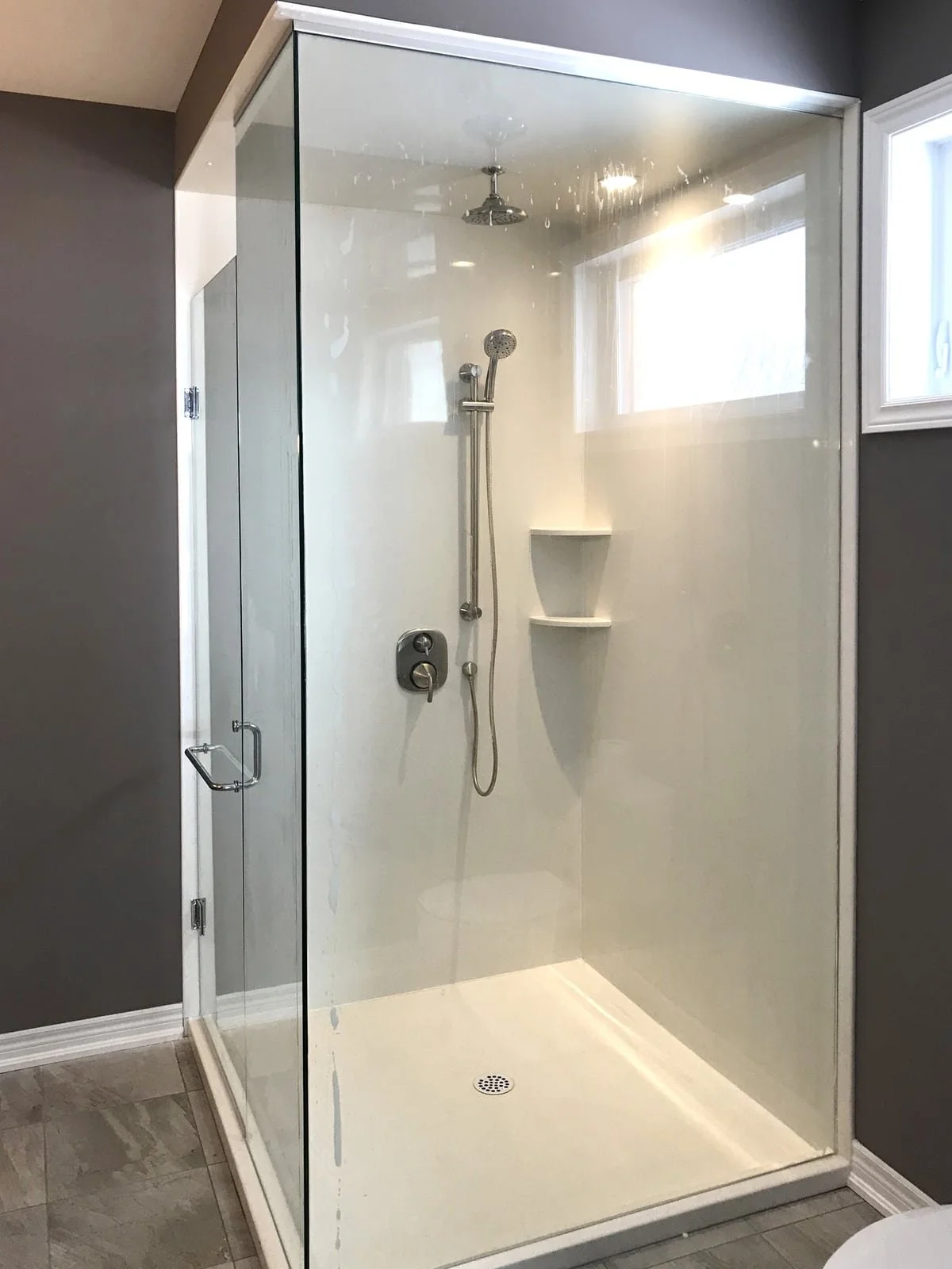 A glass-enclosed shower with gray walls, a window, a overhead showerhead, a handheld showerhead, a shelf, and a drain, with tile flooring.