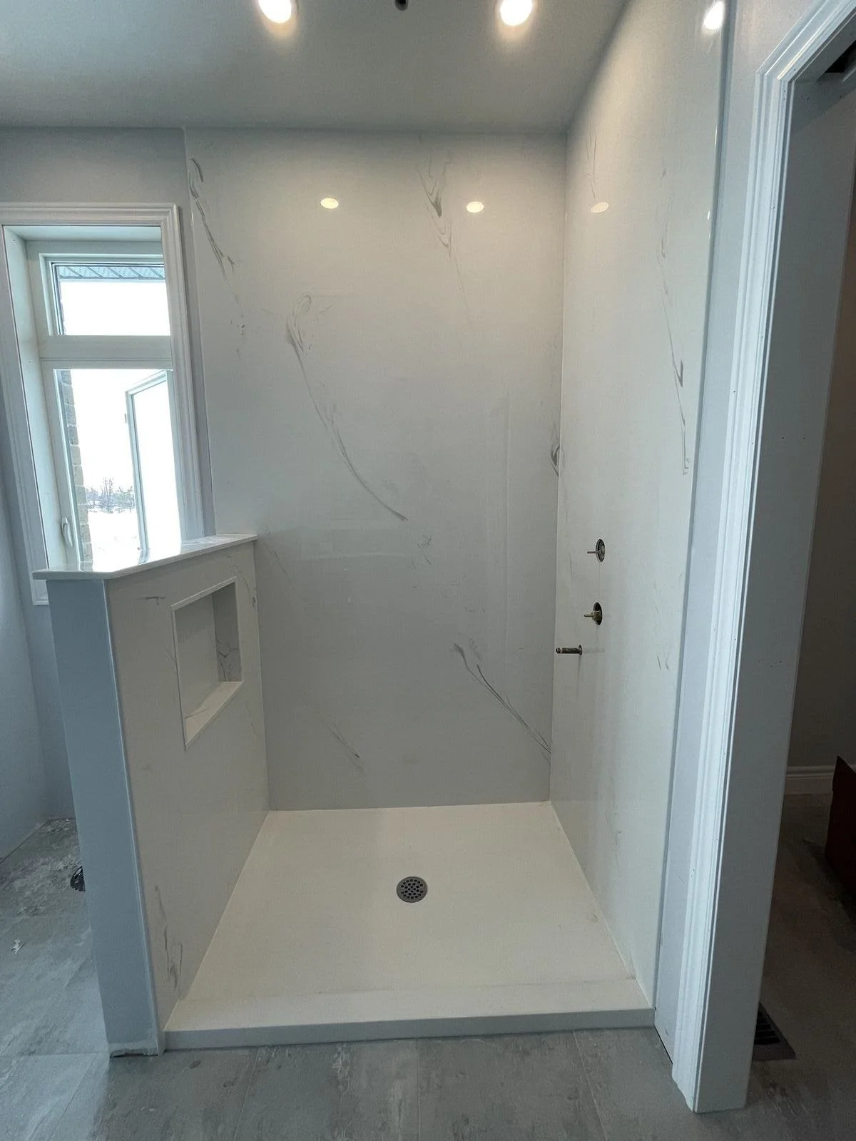 Marble Tradition, bathroom renovation in Kitchener-Waterloo & Cambridge, Ontario. Construction of a shower stall with marble walls, a drain in the floor, and plumbing fixtures installed, with an open window to the left.