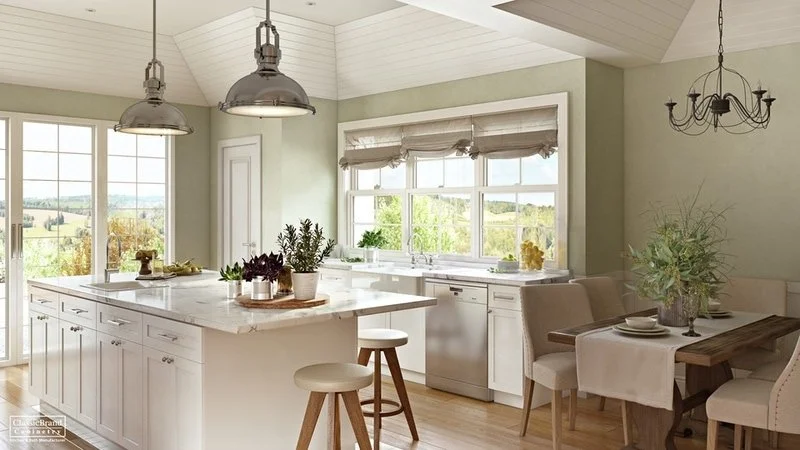 Bright kitchen with large windows, white cabinetry, central island, two hanging pendant lights, light-colored walls, and a dining area with a wooden table and beige chairs.