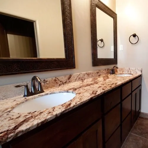 Bathroom with double sink granite countertop, dark wood cabinets, two framed mirrors, two towel rings, and tiled floor.