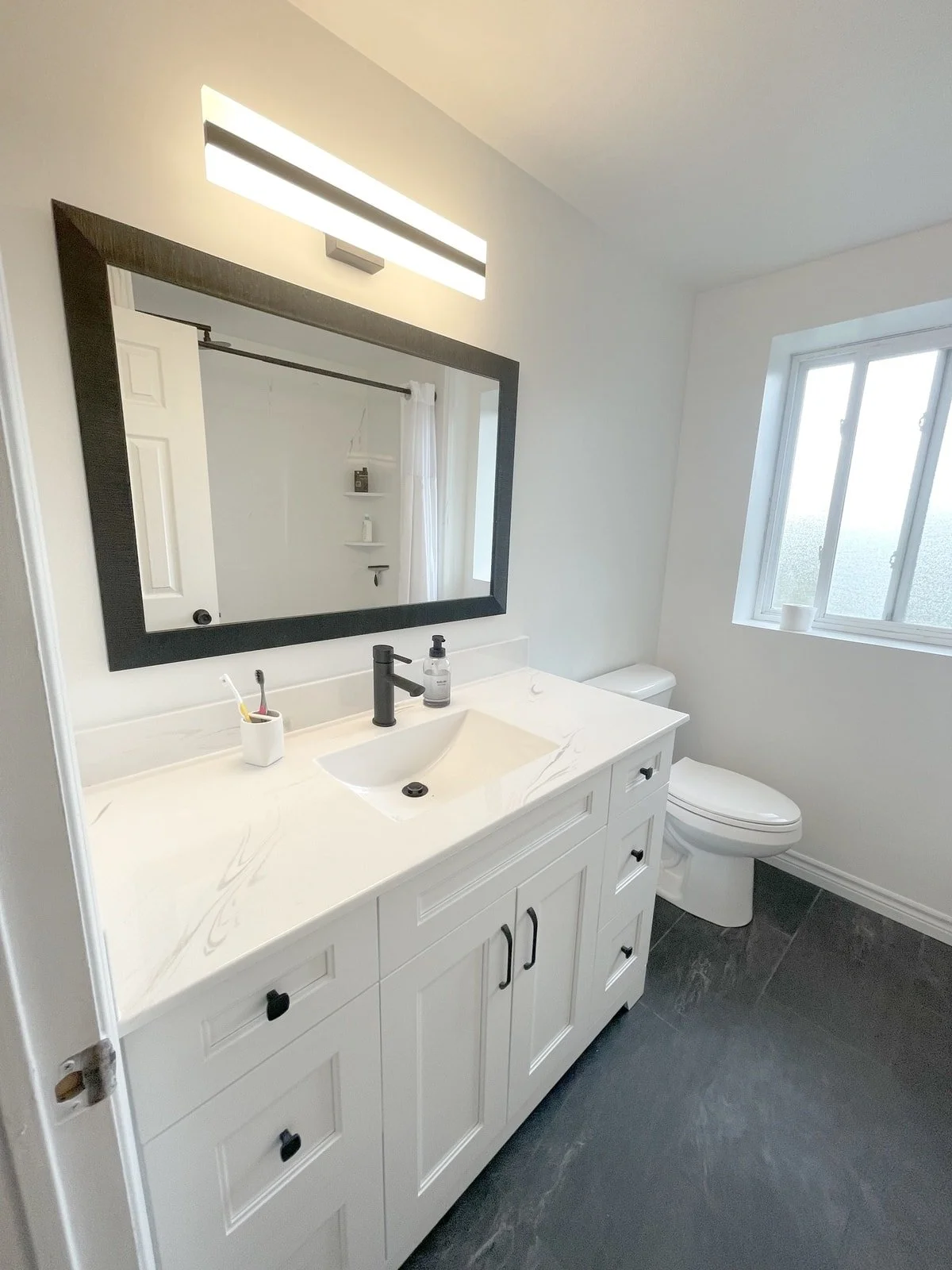 Modern bathroom with white vanity, black faucet, large mirror, light fixture, window, and toilet.