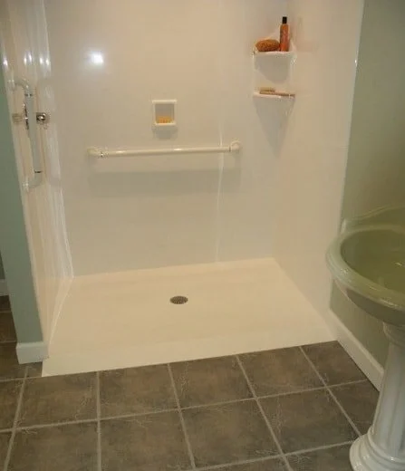 Empty shower area with a grab bar, a small wall-mounted shelf with some items, and a drain on the floor, adjacent to a toilet.