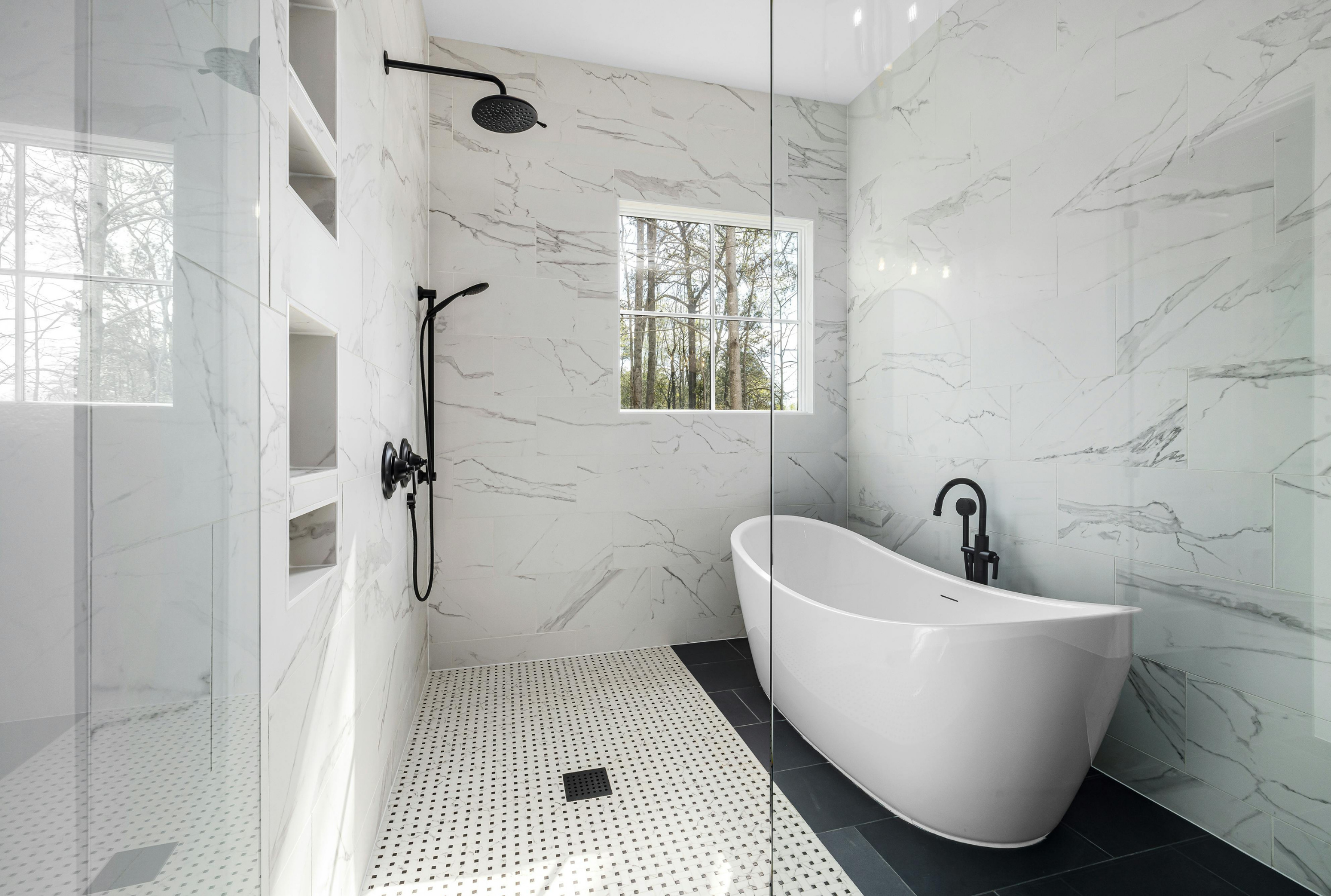 Marble Tradition, bathroom renovation in Kitchener-Waterloo & Cambridge, Ontario. A modern bathroom with marble walls, a black rainfall showerhead, a handheld shower, a window with trees outside, and a freestanding bathtub.