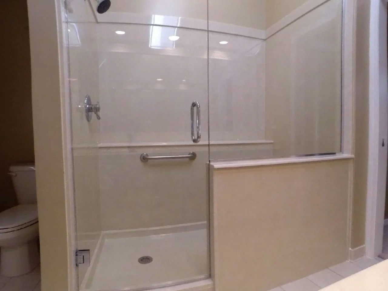 Accessible shower stall with glass doors, grab bar, and a hand-held shower head, adjacent to a toilet in a bathroom.