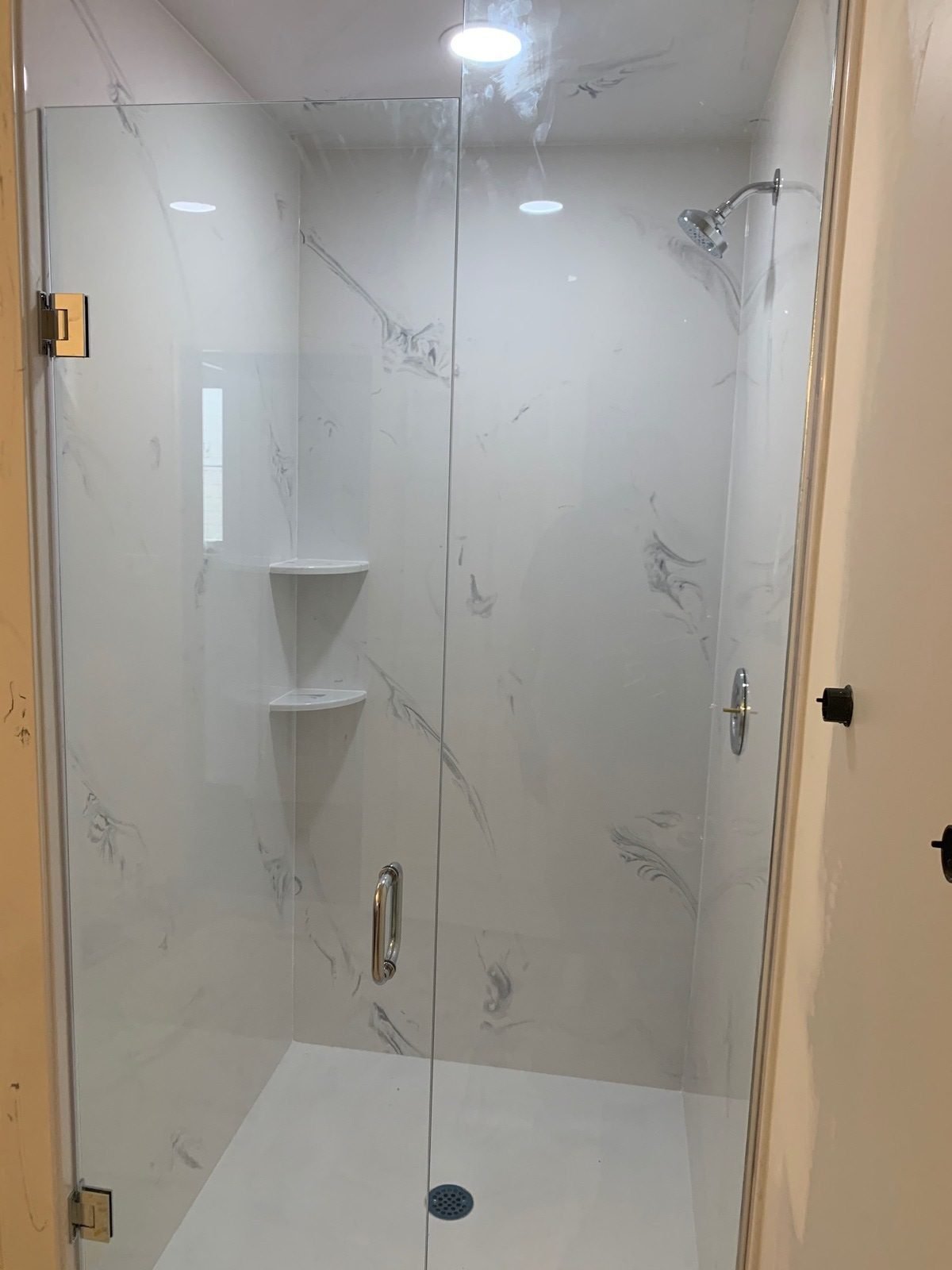 A modern glass-enclosed shower with white marble walls and built-in corner shelves.