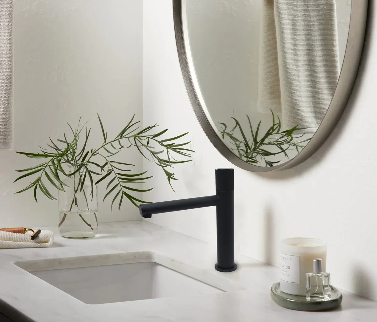 Bathroom sink with black faucet, white countertop, mirror, and decorative items including a glass vase with green leaves, scented candle, and perfume