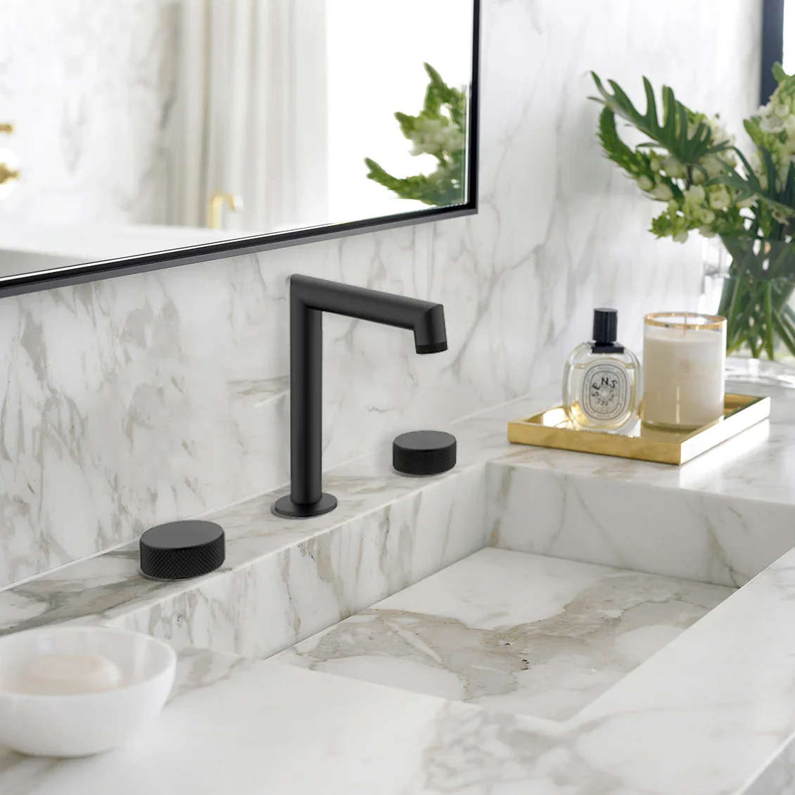 A modern bathroom sink with a black faucet, decorative black knobs, a mirror, and a tray with perfume, a candle, and green plants in the background.