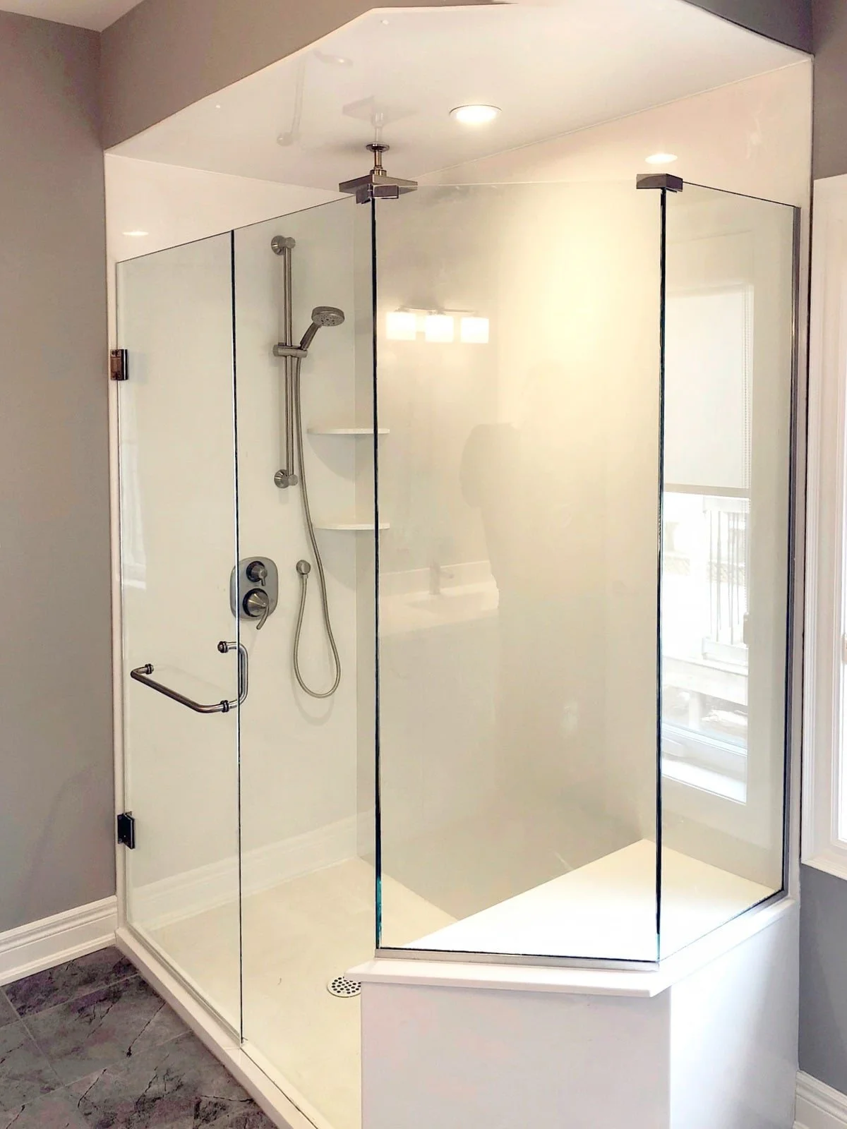 A modern glass shower enclosure with a stainless steel handheld showerhead and a wall-mounted control panel, featuring built-in corner shelves for storage, situated in a bathroom with gray tiled flooring and a window with blinds.