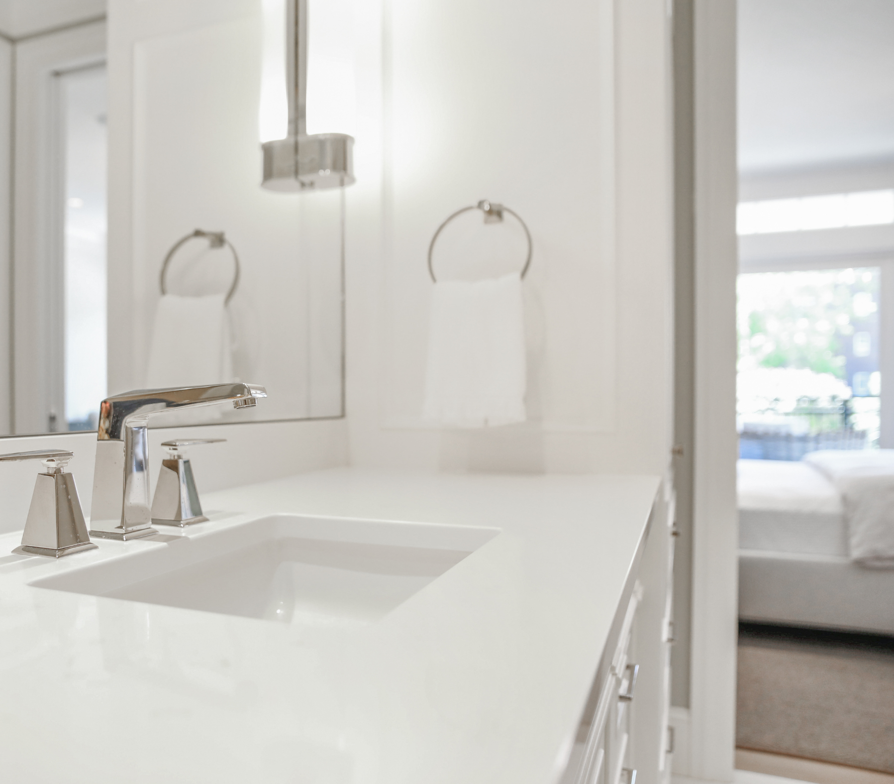 Marble Tradition, bathroom renovation. Close-up of a white bathroom vanity with a white sink, silver faucet, and cabinet drawers, reflected in a mirror with towel rings on a white wall, and a window with blinds letting in natural light.