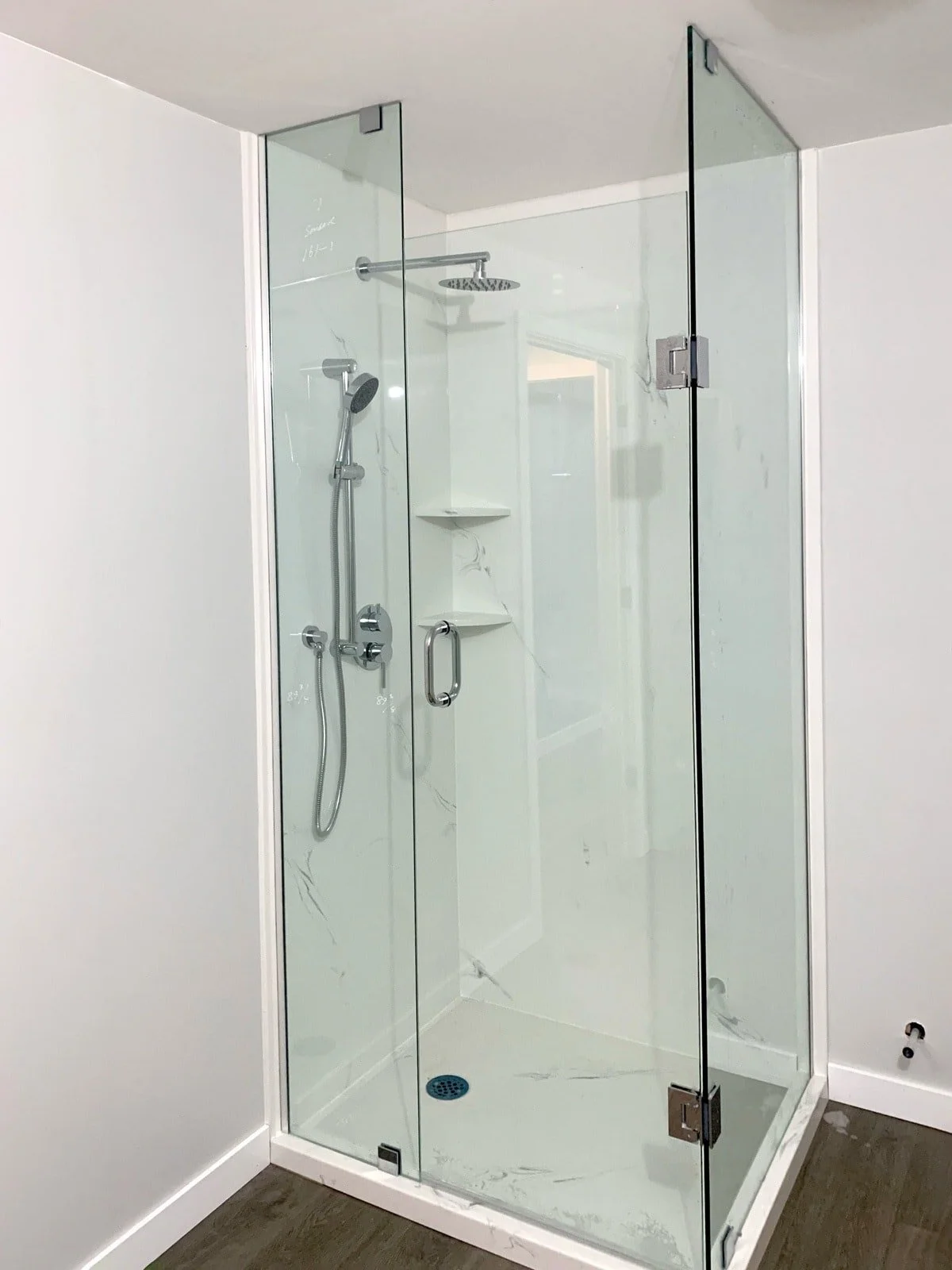 A modern glass shower enclosure with a metal handle, dual shower heads, and built-in corner shelves, surrounded by white walls and dark flooring.