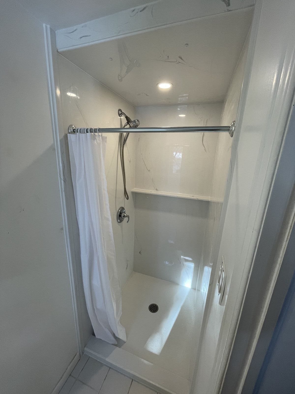 Marble Tradition, bathroom renovation in Kitchener-Waterloo & Cambridge, Ontario. A small shower stall with a white curtain, a handheld showerhead, a built-in shelf, and a drain on the floor.