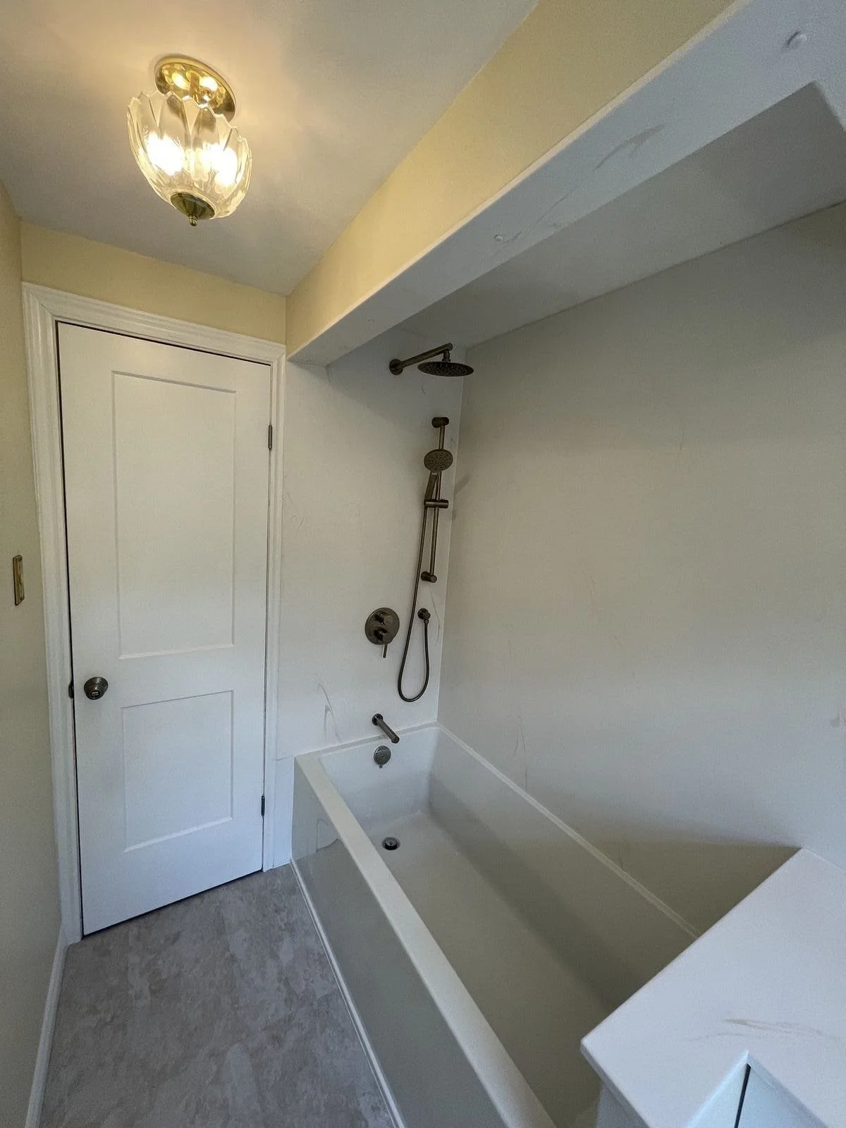 A small bathroom with a white bathtub, a showerhead on a wall, a closed white door, and a ceiling light fixture.
