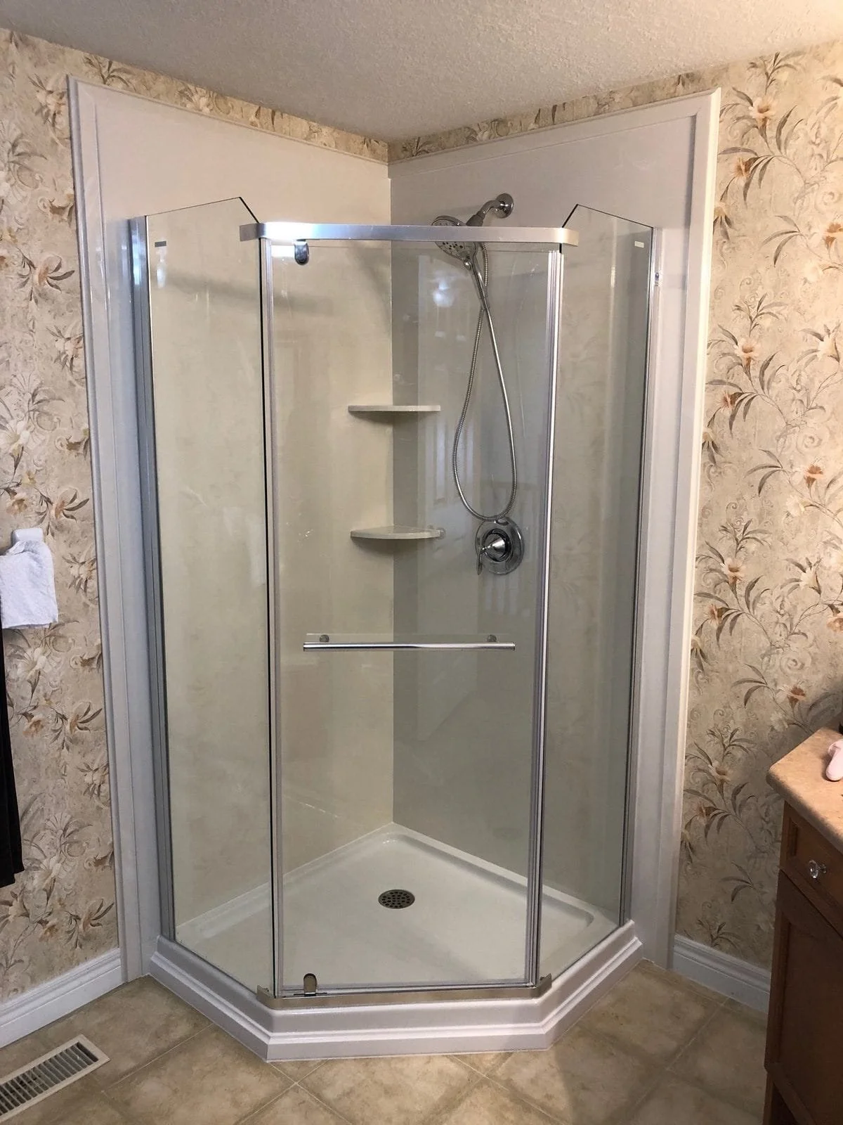 A corner shower with glass doors, beige tile walls, built-in shelves, and a showerhead, in a bathroom with floral wallpaper.