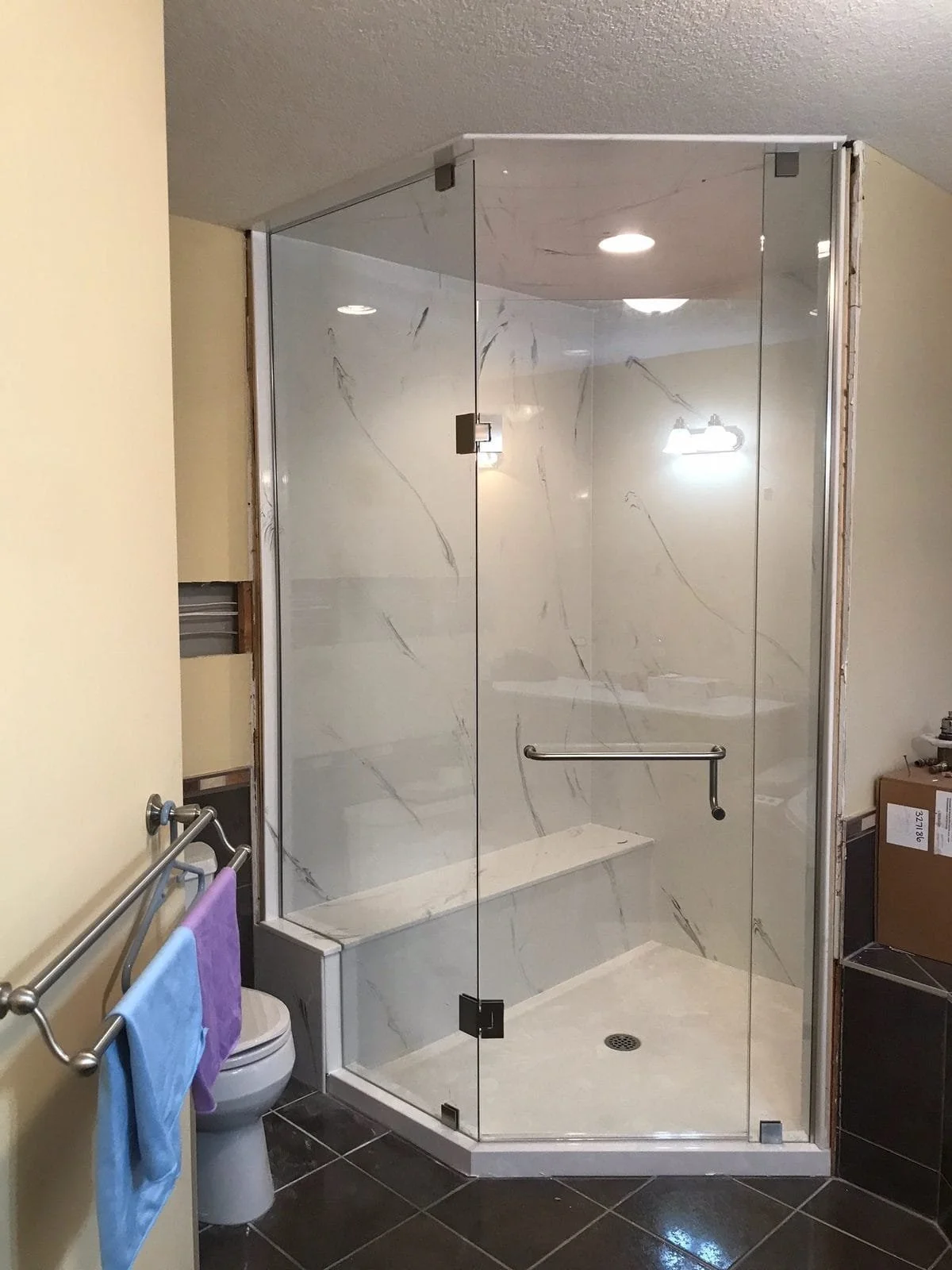 A bathroom with beige walls and dark tiled flooring, featuring a glass shower enclosure with marble-patterned walls and a built-in bench, a towel bar with towels, and a partial view of a toilet and boxes.