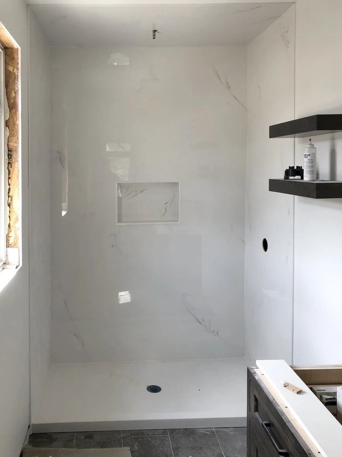 A newly constructed bathroom shower with white marble walls, a square drain on the floor, an opening for a showerhead, and two black floating shelves with toiletries on the right wall. There's a window on the left wall, and construction materials are