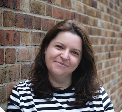 Woman with shoulder-length brown hair and a striped black and white shirt standing outdoors in front of a brick wall, smiling at the camera.