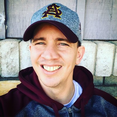 Young man smiling outdoors, wearing a baseball cap with a logo, a maroon hoodie, and a jacket, with a wooden fence and concrete blocks in the background.
