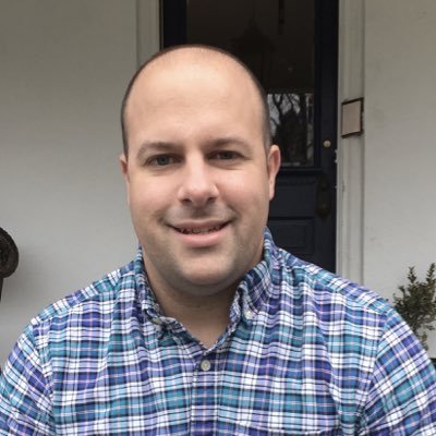 Portrait of a man with a shaved head, smiling, wearing a blue plaid shirt, standing outside a house.