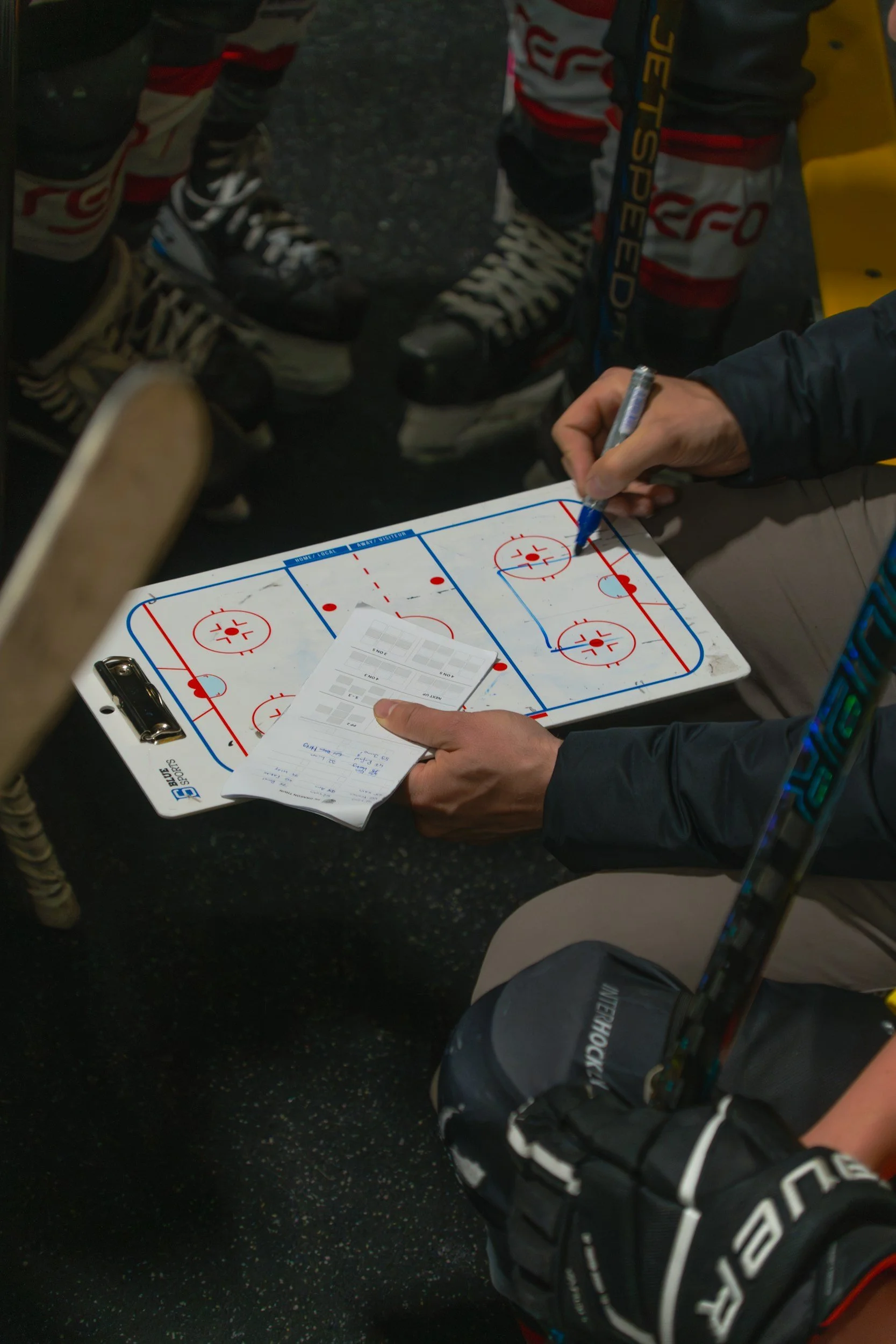 An ice hockey game in progress, with players wearing skates and helmets, and a person signing a whiteboard with hockey rink markings using a blue marker.