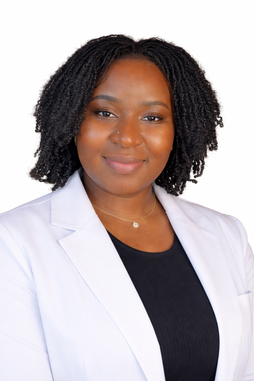 A woman with curly black hair wearing a white coat, black top, and gold jewelry, smiling against a white background.