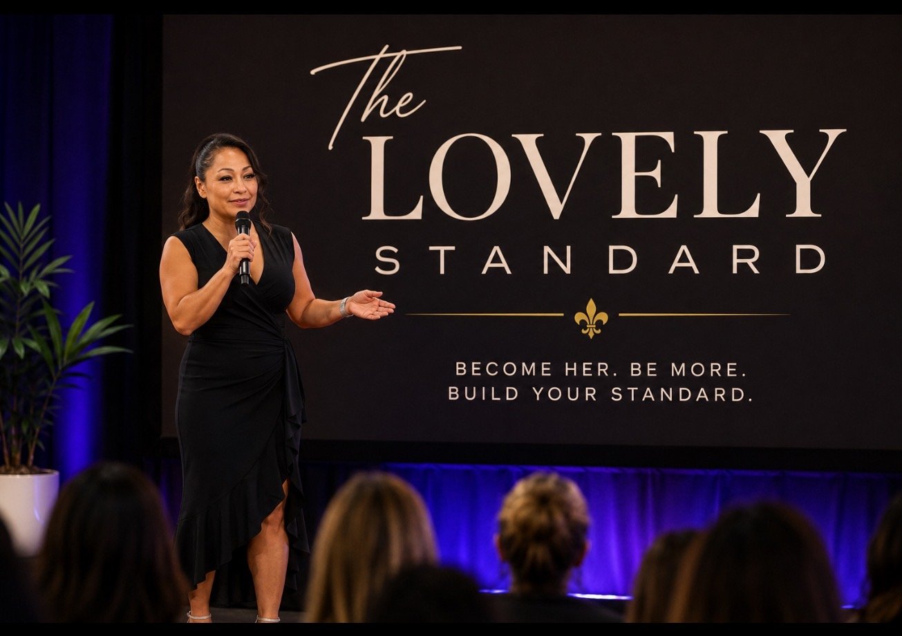 Woman in a black dress speaking into a microphone on stage at a conference, with a large screen behind her displaying the words "The Lovely Standard" and the slogan "Become her. Be more. Build your standard."