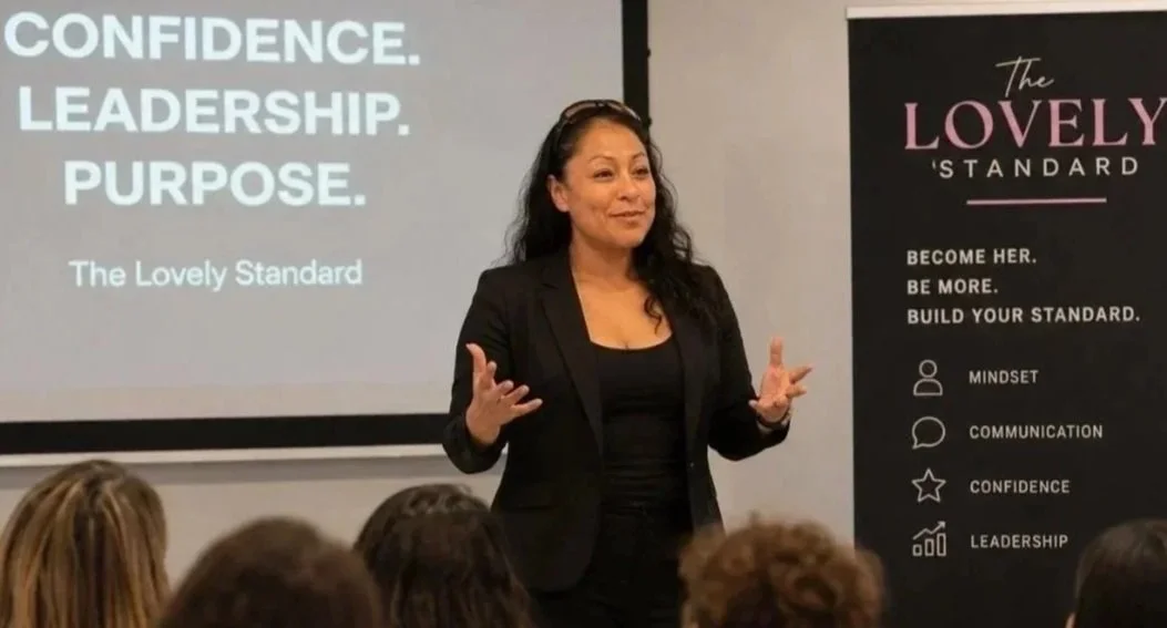 A woman giving a presentation in front of a screen and a banner that reads 'The Lovely Standard' with words like mindset, communication, confidence, and leadership listed.