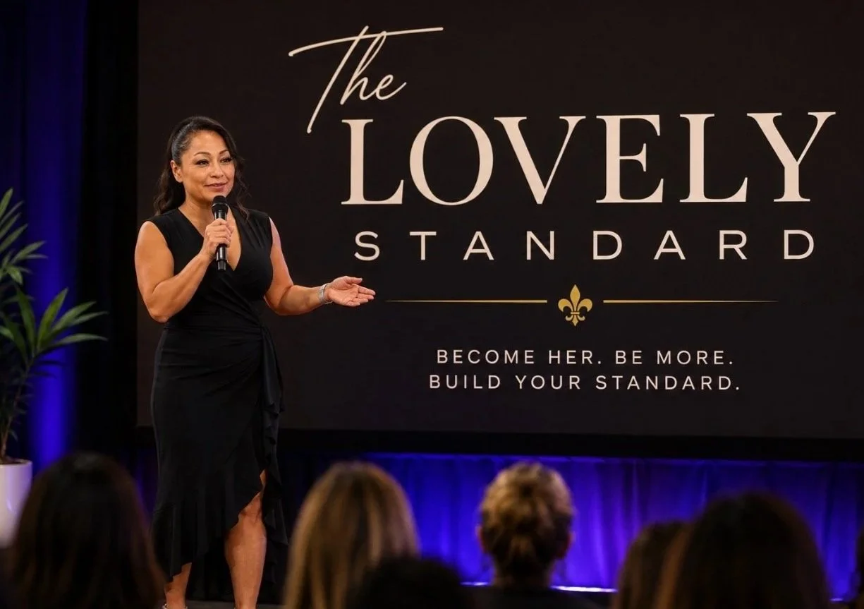 A woman in a black dress speaking into a microphone on stage at an event called "The Lovely Standard" with a large black and gold sign behind her that says "Become her. Be more. Build your standard." Audience members are visible in the foreground.
