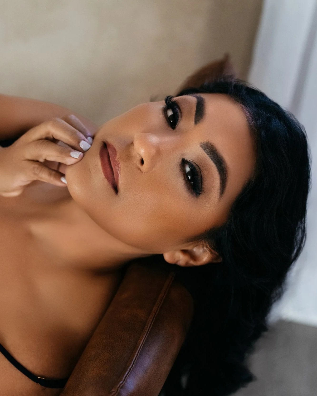 A woman with dark hair and makeup, lying on a brown surface with her hand near her chin, looking at the camera.