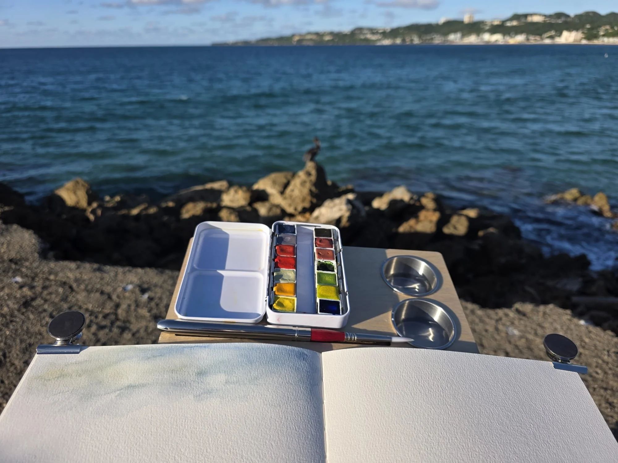 A watercolor palette with various colors, a jar of water, and a paintbrush on a white table at a rocky shoreline. In the background, the ocean with small waves and a distant hilly coastline with buildings can be seen.
