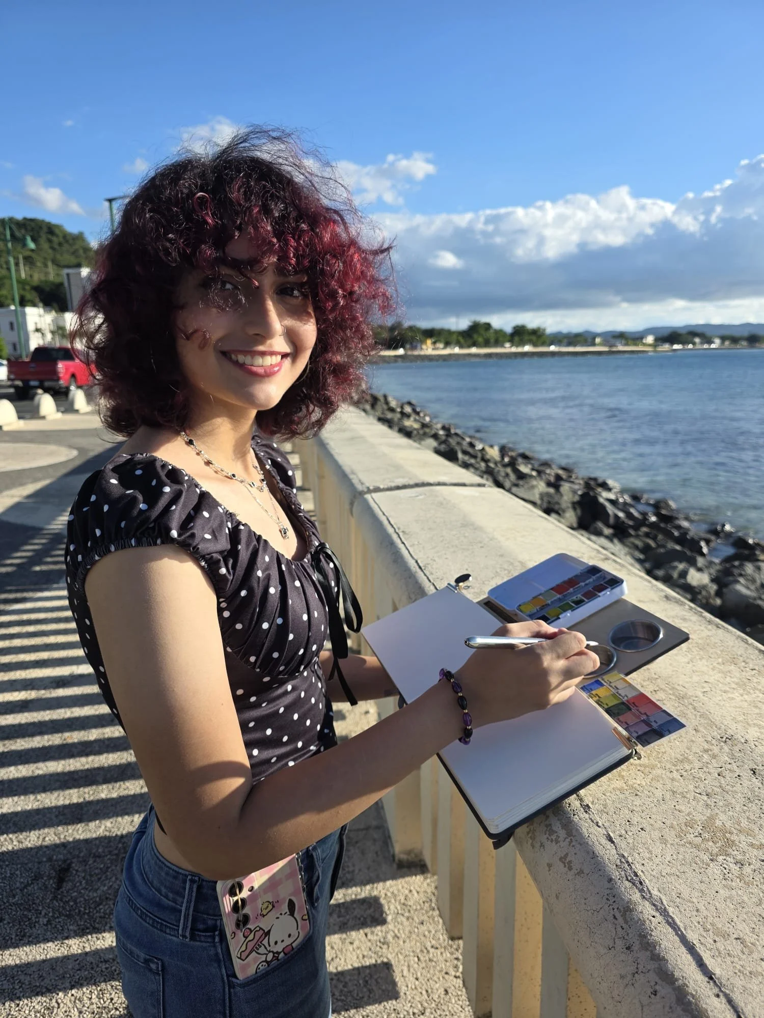 Woman with curly red hair and polka dot top painting by the water with watercolor paints and a sketchbook.