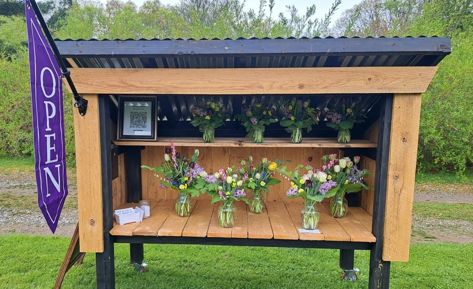 A small flower booth with a purple 'OPEN' flag, displaying arranged flower bouquets in glass jars on two shelves made of wood, set against a green outdoor background with trees.