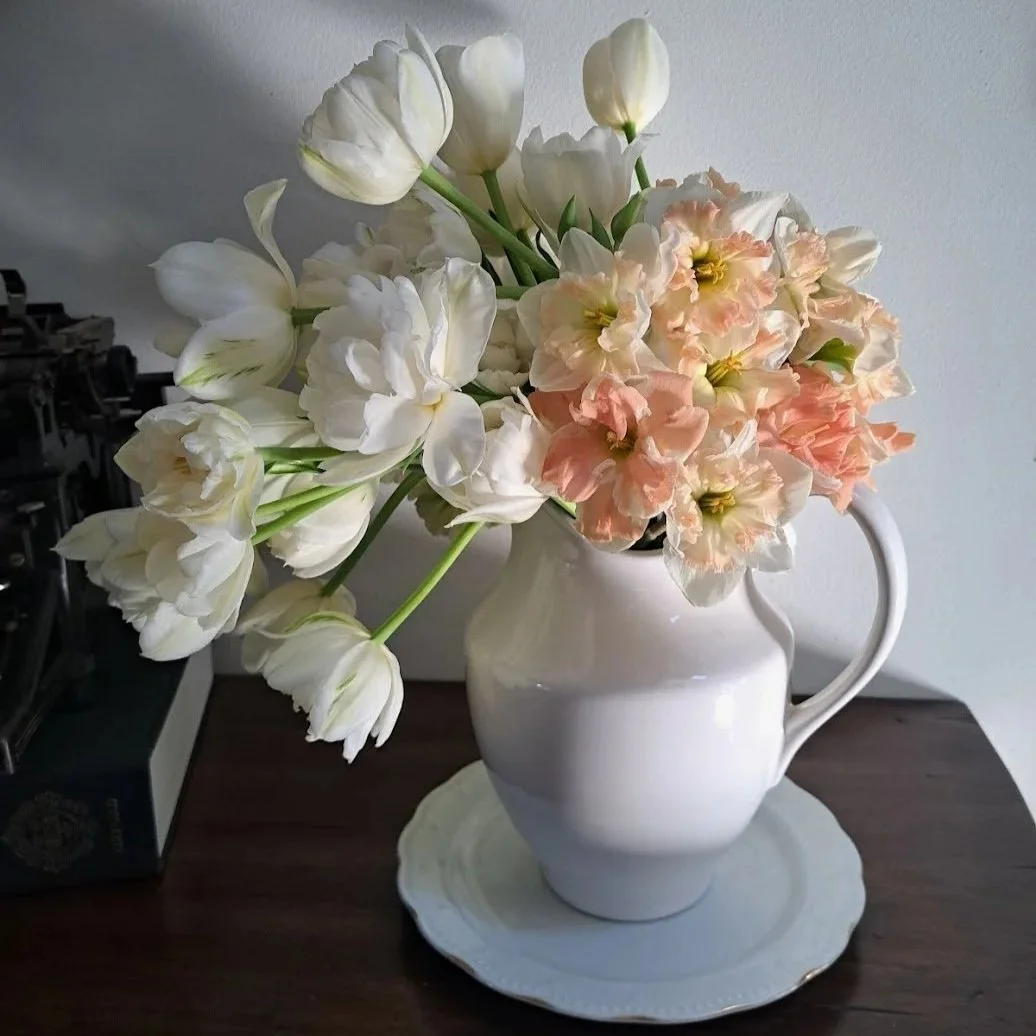 A white ceramic pitcher filled with white and light pink flowers, placed on a matching white tray on a wooden surface.