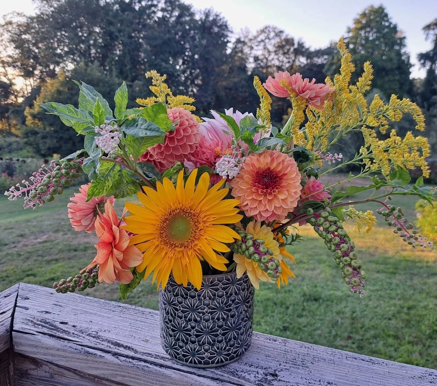 A colorful bouquet of flowers in a geometric black and white vase, placed on a wooden surface outdoors with a blurred green landscape and trees in the background.