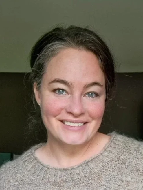 Close-up of a smiling woman with light skin, blue eyes, brown hair with gray streaks, and wearing a beige sweater.