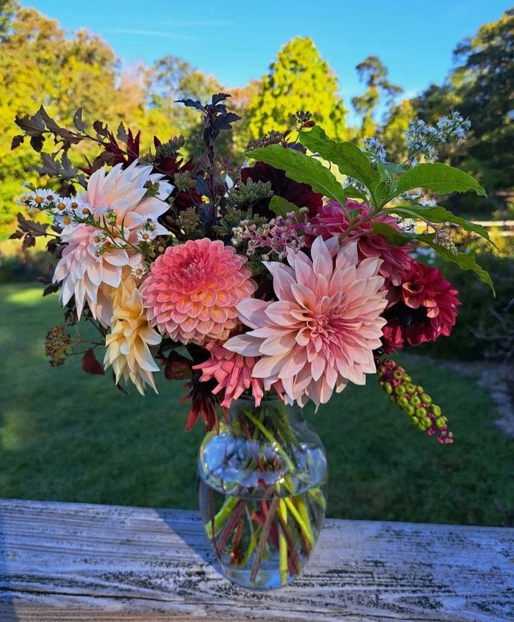 A glass vase filled with pink, white, and cream-colored dahlias, small daisies, and greenery, set outdoors on a weathered wooden surface with a background of green trees and blue sky.