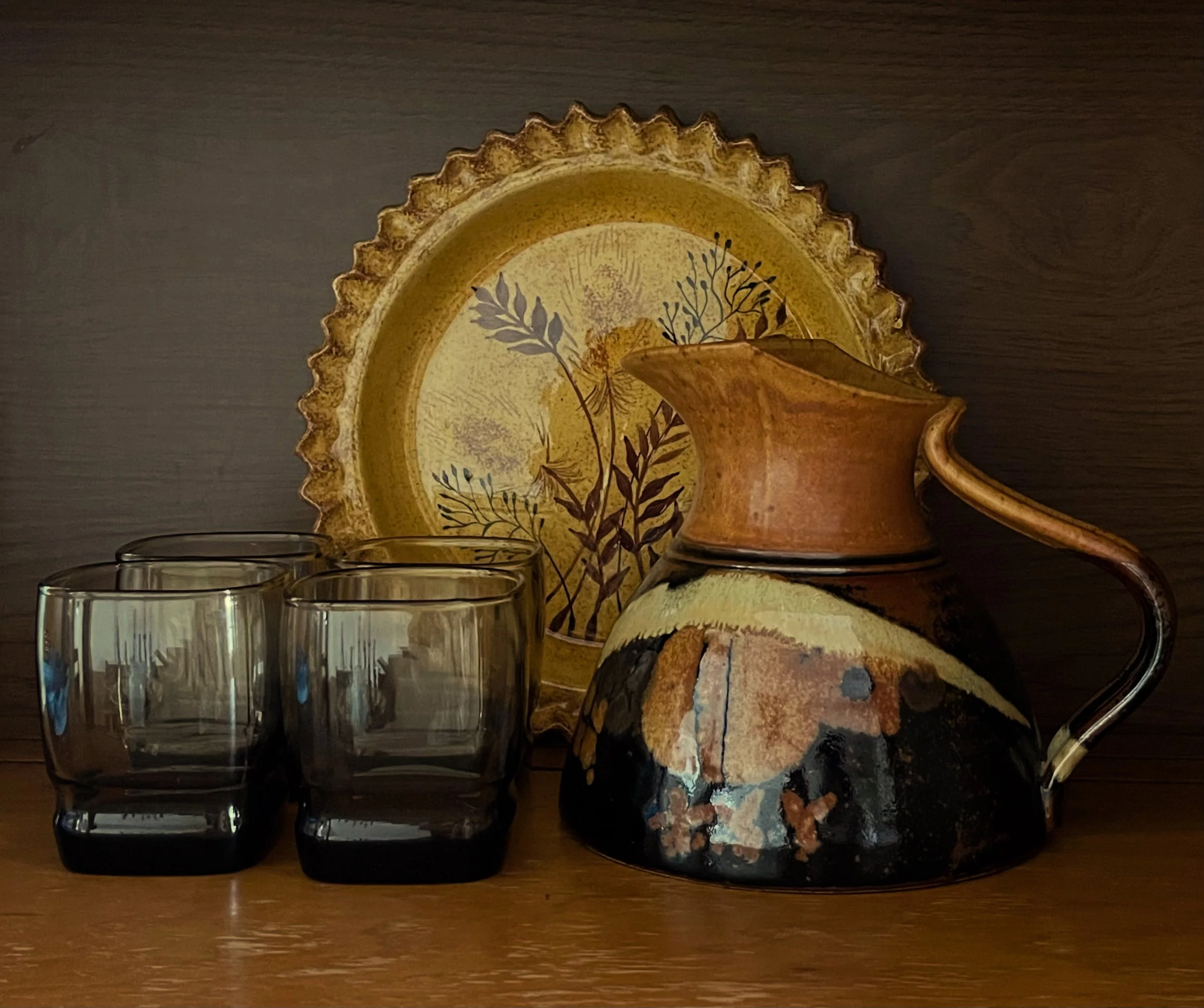 A decorative ceramic plate with botanical artwork, placed against a dark background, partly behind a brown vase. Three transparent glasses and a small ceramic piece are at the bottom, all on a wooden surface.